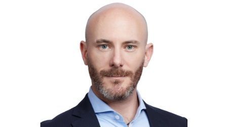 Bald man with a trimmed beard and light blue shirt, wearing a navy blazer, looking directly at the camera.