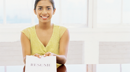 Woman smiling, holding a resume. Wearing yellow top, sitting at a table.