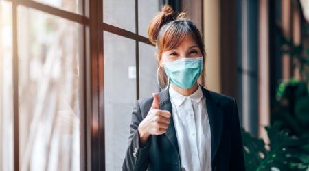 Woman in black blazer and face mask giving thumbs up near a window.