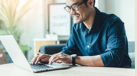 Man in glasses smiles, typing on laptop at a table, wearing a blue shirt, bright setting.