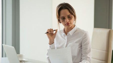 Woman in glasses reading documents in an office, holding pen, white shirt.