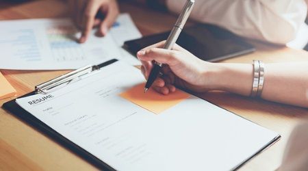 Person writing on a resume, possibly reviewing or editing it, with documents and pen on a table.