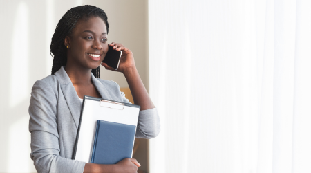 Woman in gray suit, holding files, smiles while talking on a smartphone near a bright window.