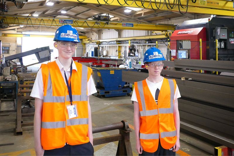 Two men wearing hard hats and orange vests are standing in a factory