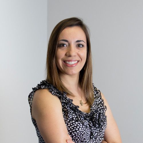 Woman with brown hair and a patterned top smiles, arms crossed. Light background.