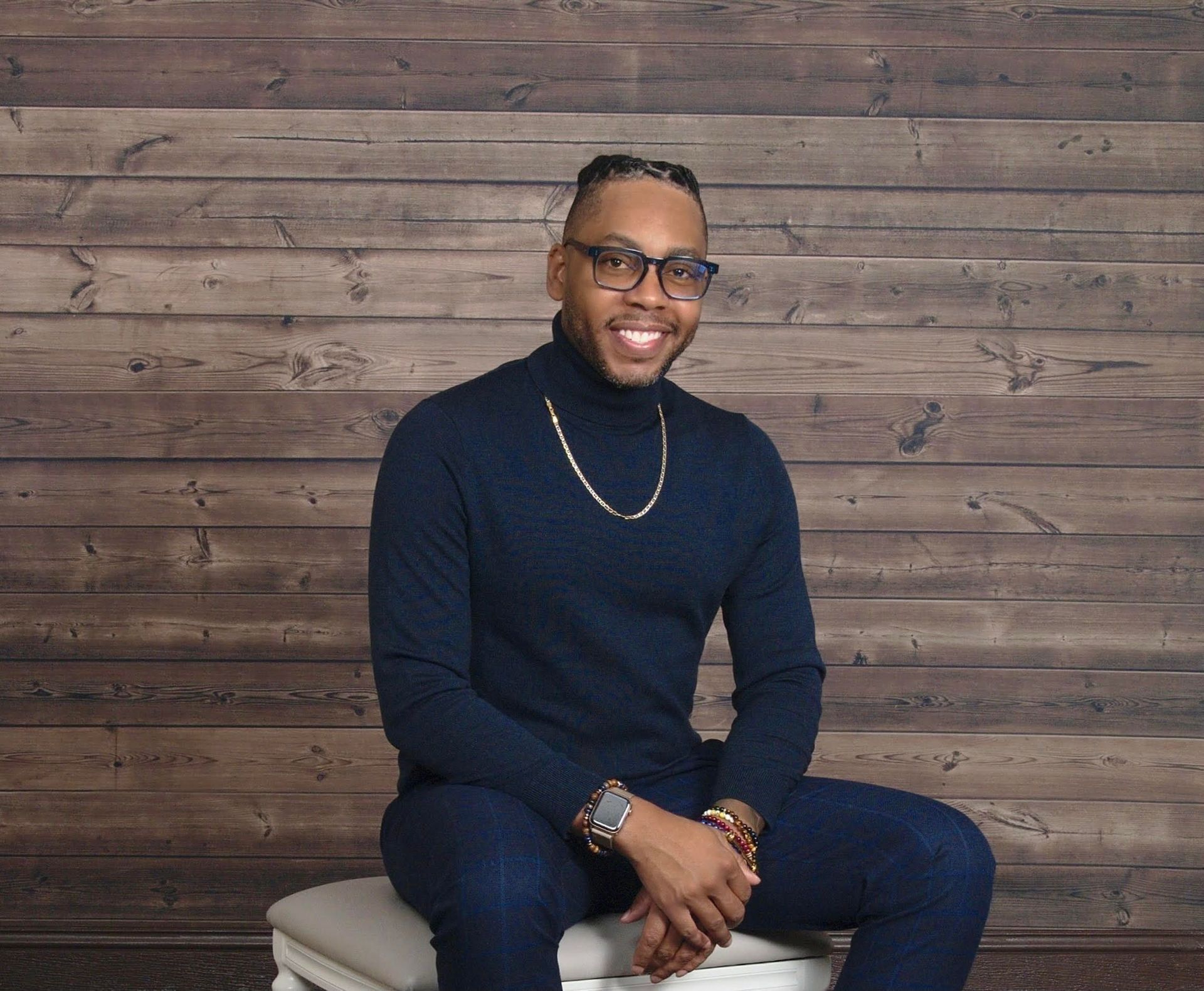 A smiling Black man with glasses in a blue turtleneck and jeans, sitting on a stool against a wood background.