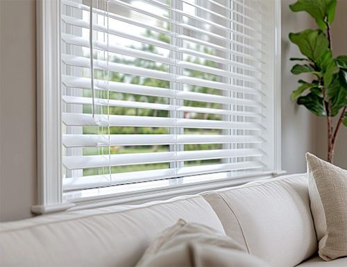 White wooden blinds on a window, partially covering the view. A white sofa sits below, with a plant to the right.