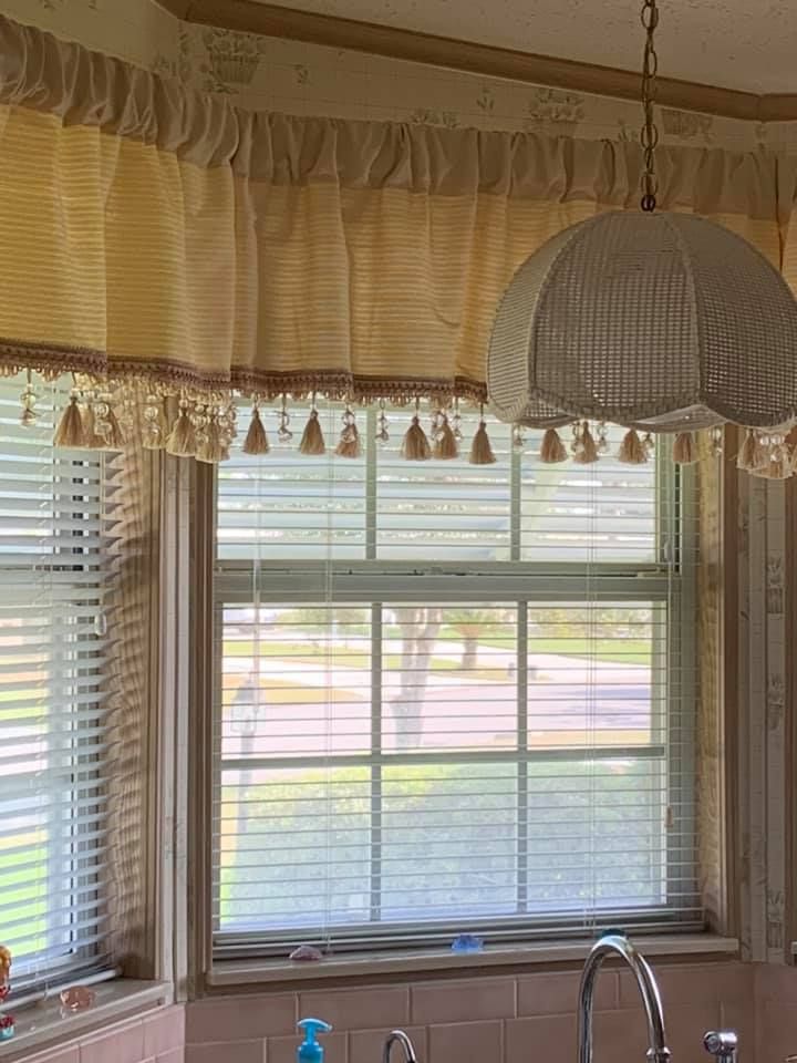 Window with blinds, a valance with tassels, and a wicker pendant lamp over a sink.