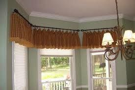 Tan pleated curtains on a curved rod above a bay window, with a chandelier hanging nearby.
