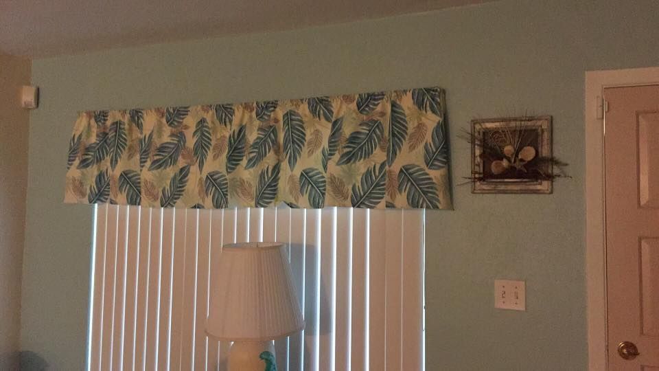Window with leaf-patterned valance, blinds, lamp, framed art, and light blue wall.