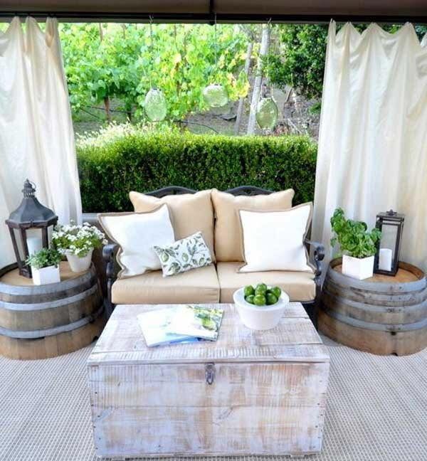 Outdoor patio with couch, barrel side tables, and distressed wood coffee table, overlooking a garden.