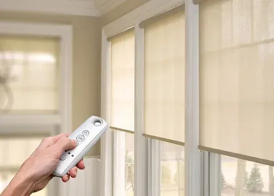 Hand holding remote controlling beige window roller shades in a bright room.