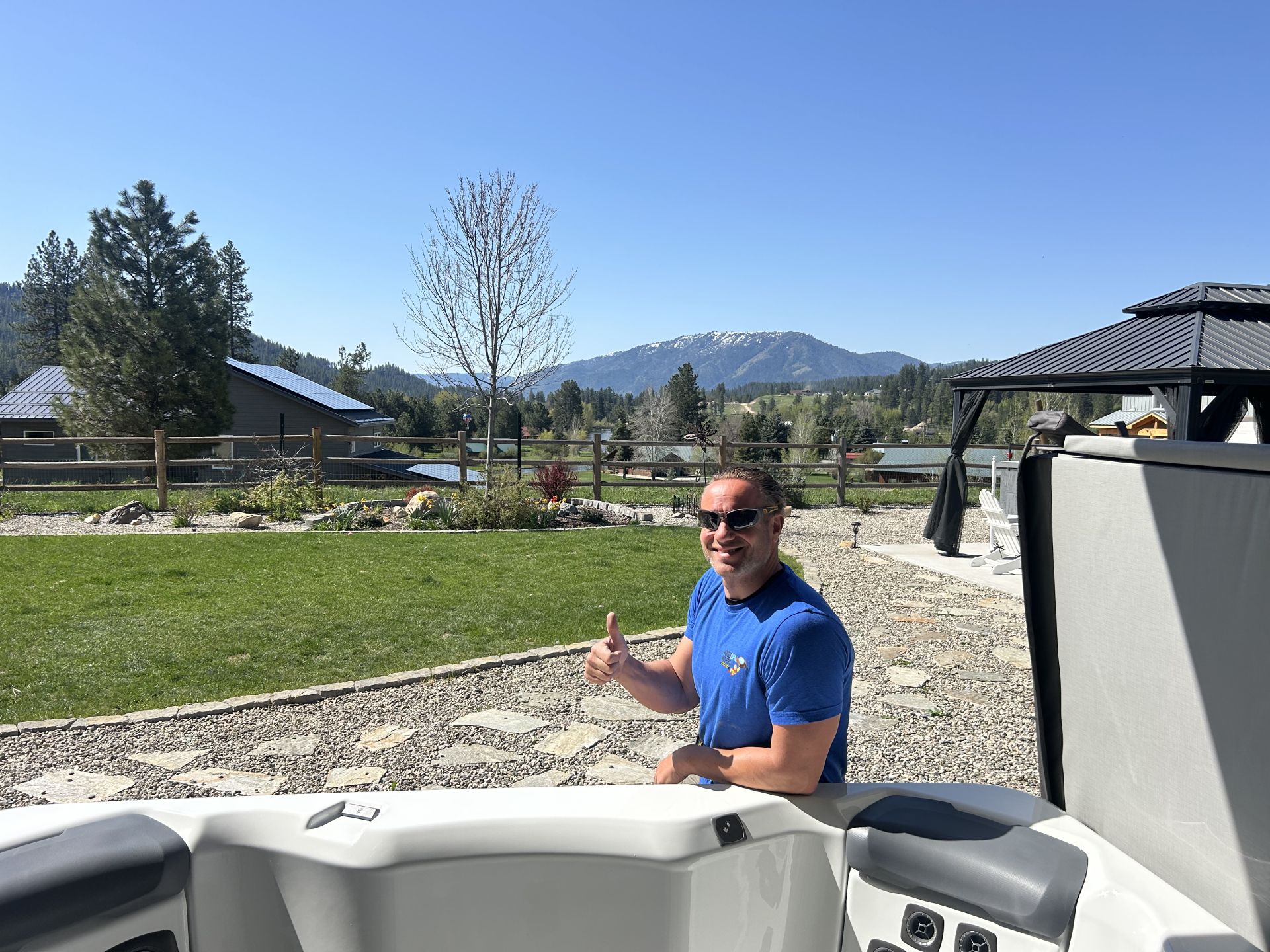 Man in blue shirt gives thumbs-up from a hot tub, sunny backyard with mountains in the background.