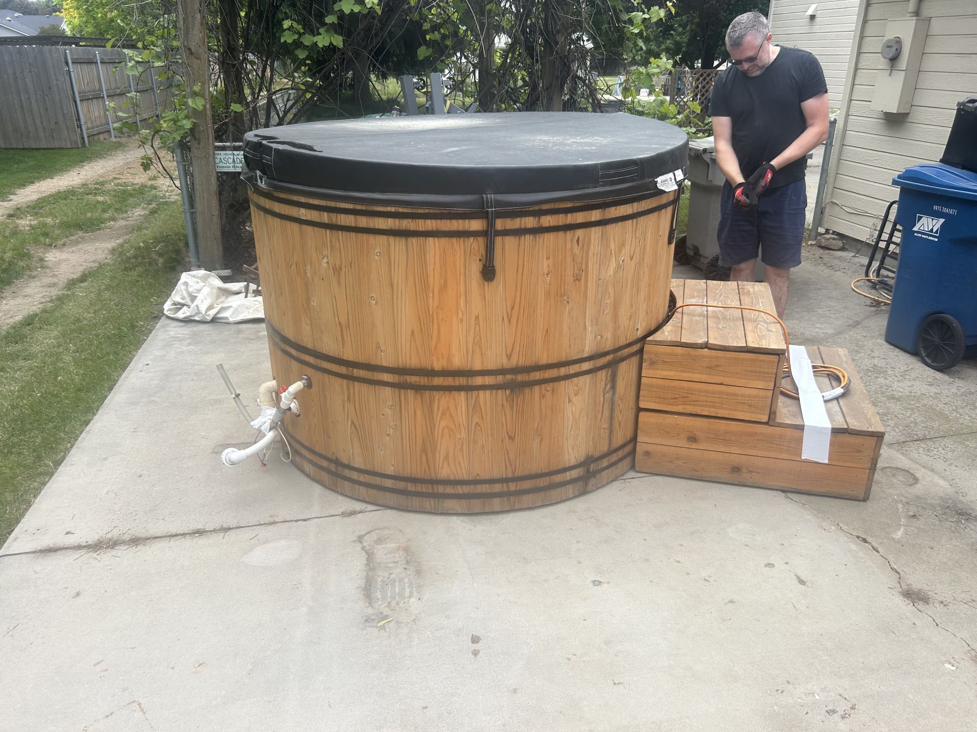Wooden hot tub with cover on a concrete patio, man standing nearby.