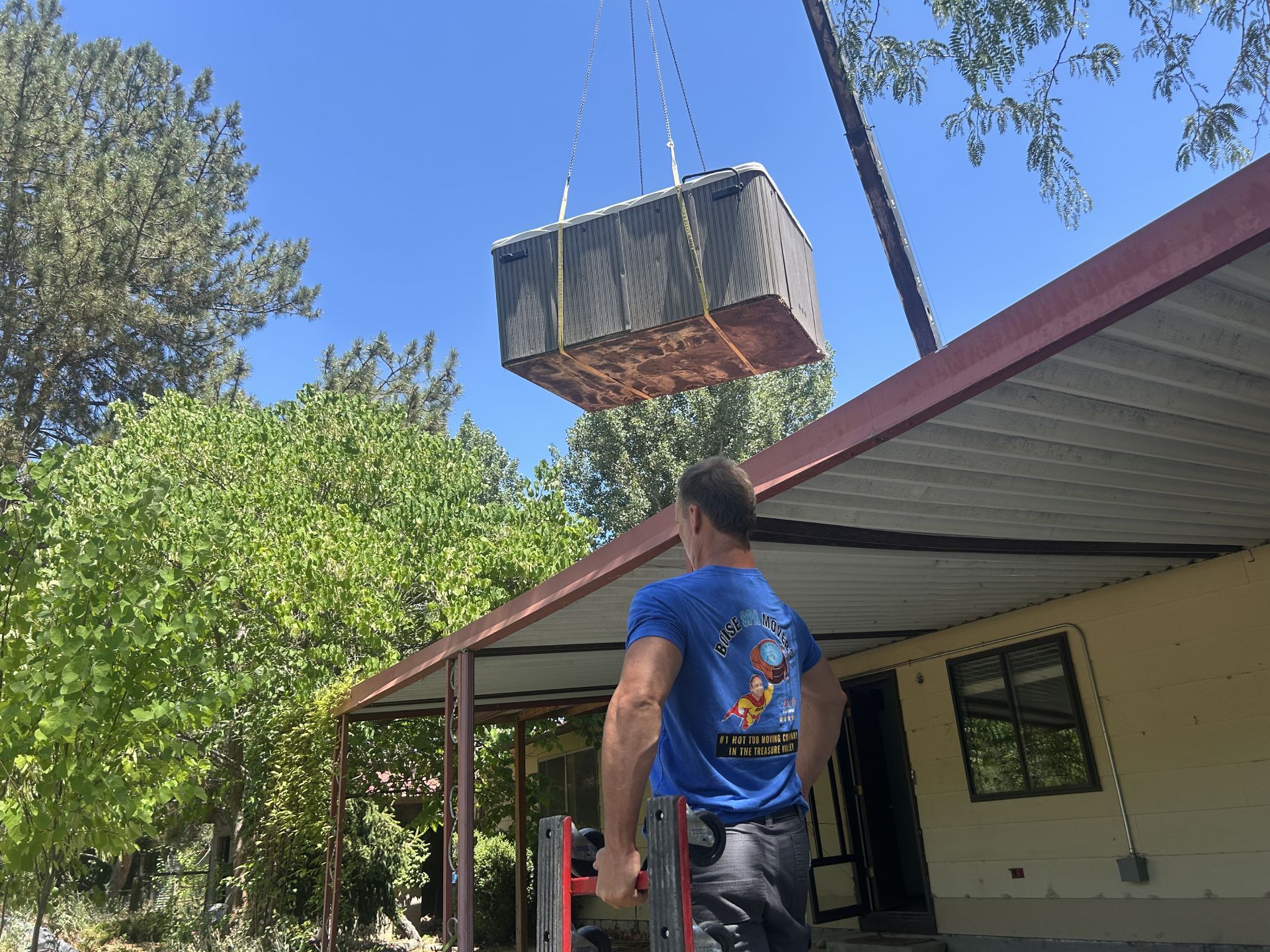 A man watches as a hot tub is lifted by a crane near a house with a red awning on a sunny day.