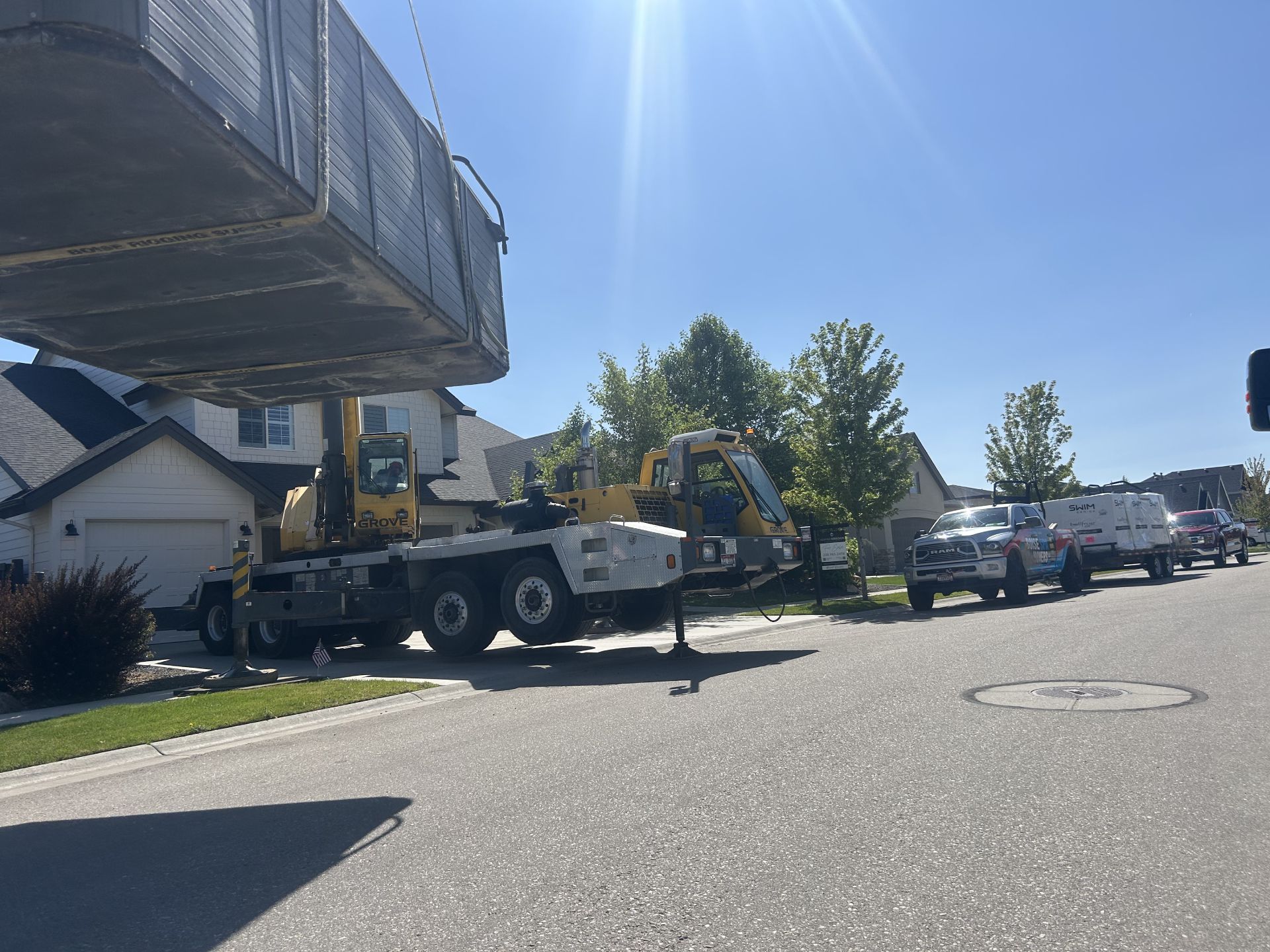 Crane lifting a large rectangular object in a residential street on a sunny day. Trucks and houses are also present.