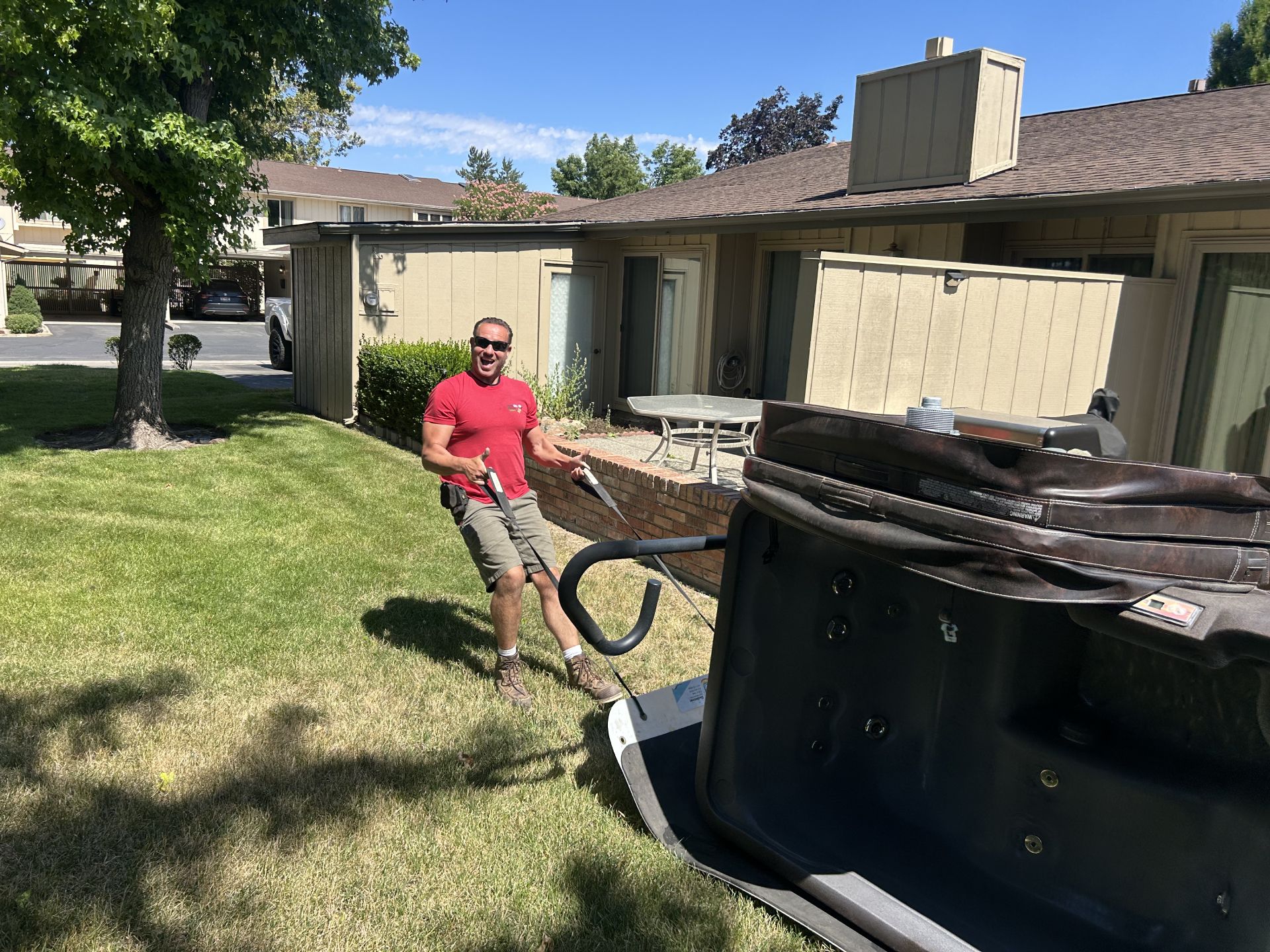 Man in a red shirt pulling a hot tub across a lawn, in front of a building.