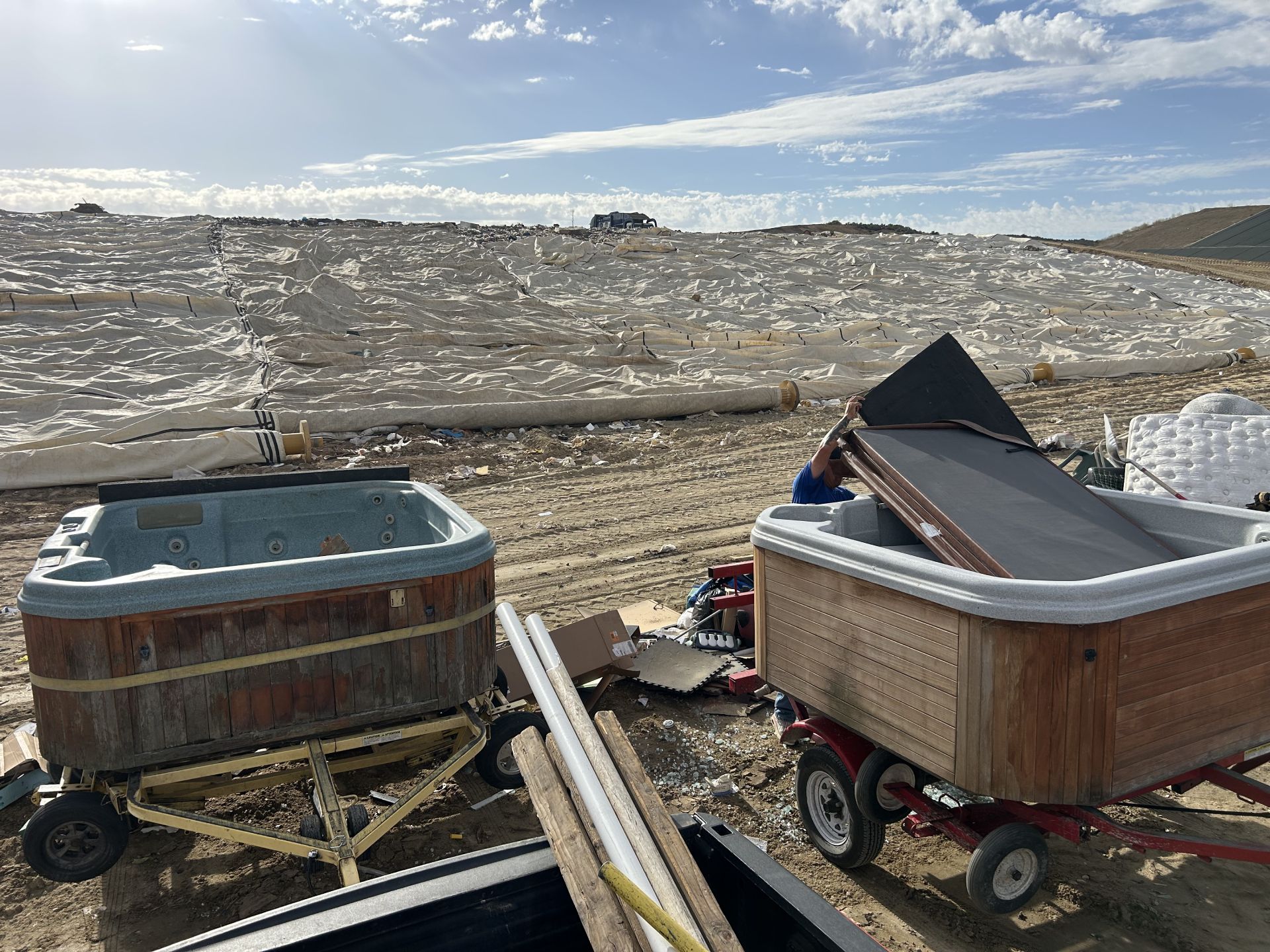 Two hot tubs on trailers sit in a field; one has its cover open. In the background, white fabric covers a hillside.