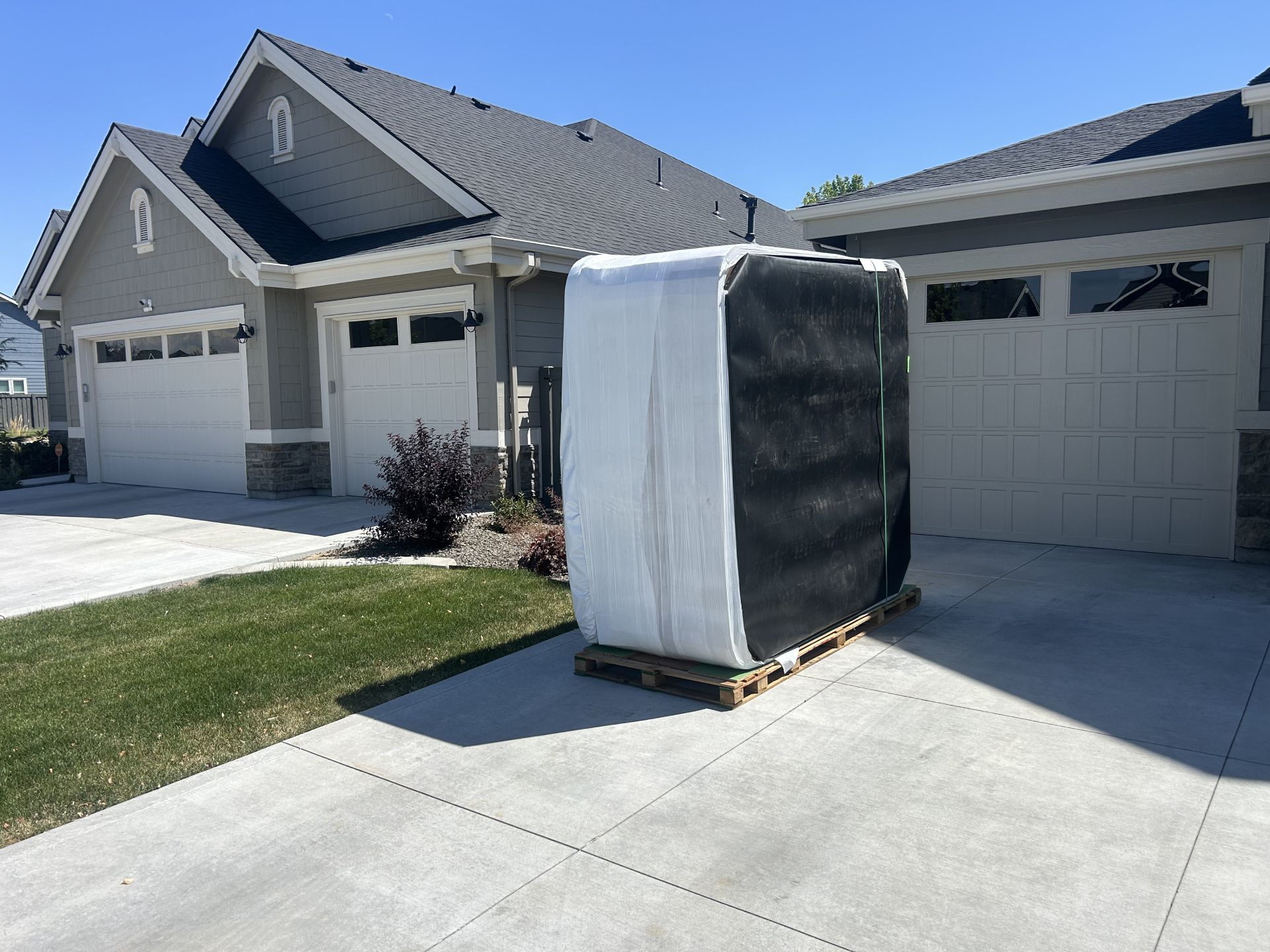 Large, wrapped object on a pallet in a driveway, in front of a house with white garage doors, under a blue sky.