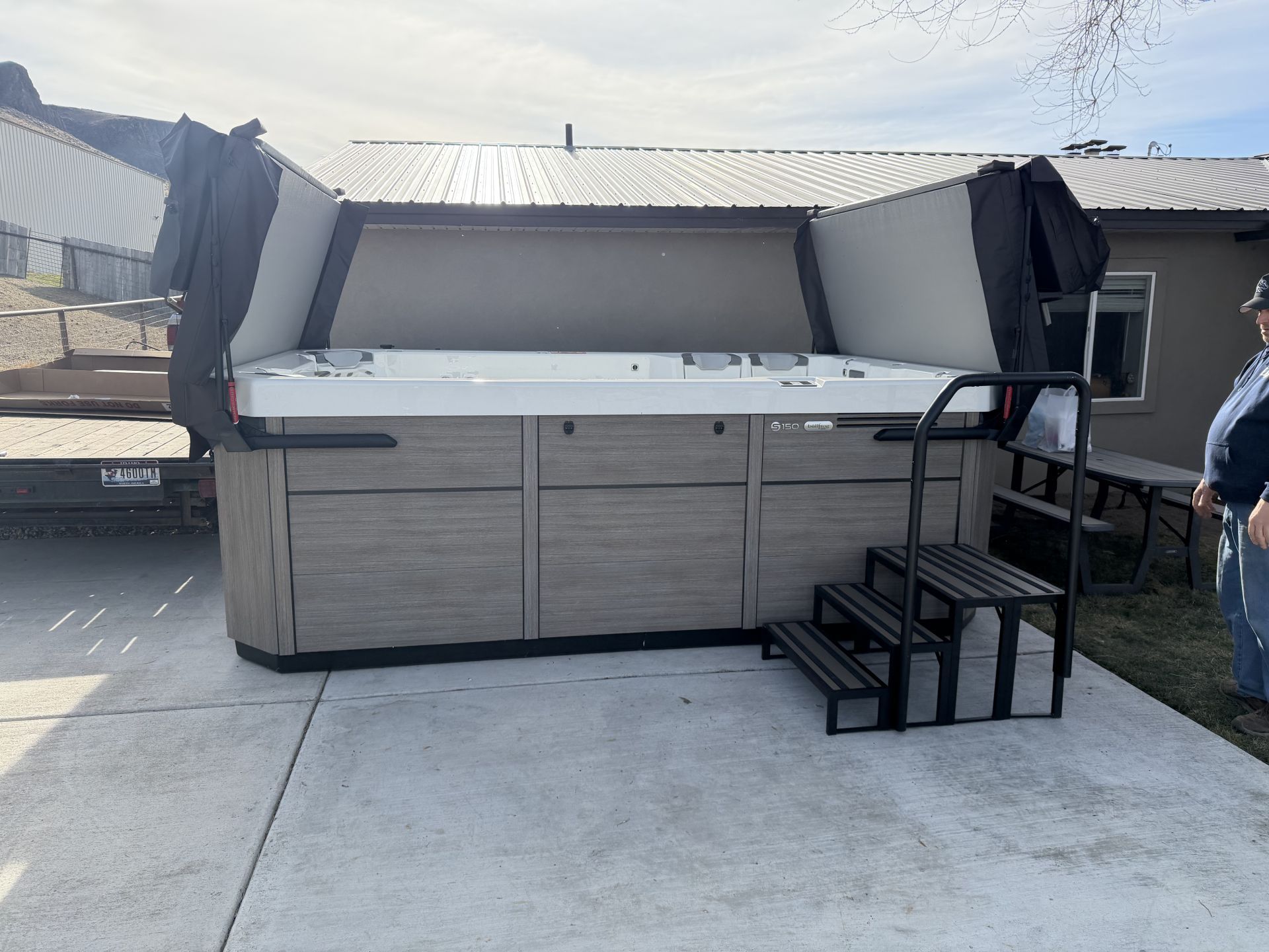 Hot tub with open cover, steps, and a person standing nearby on a concrete patio.