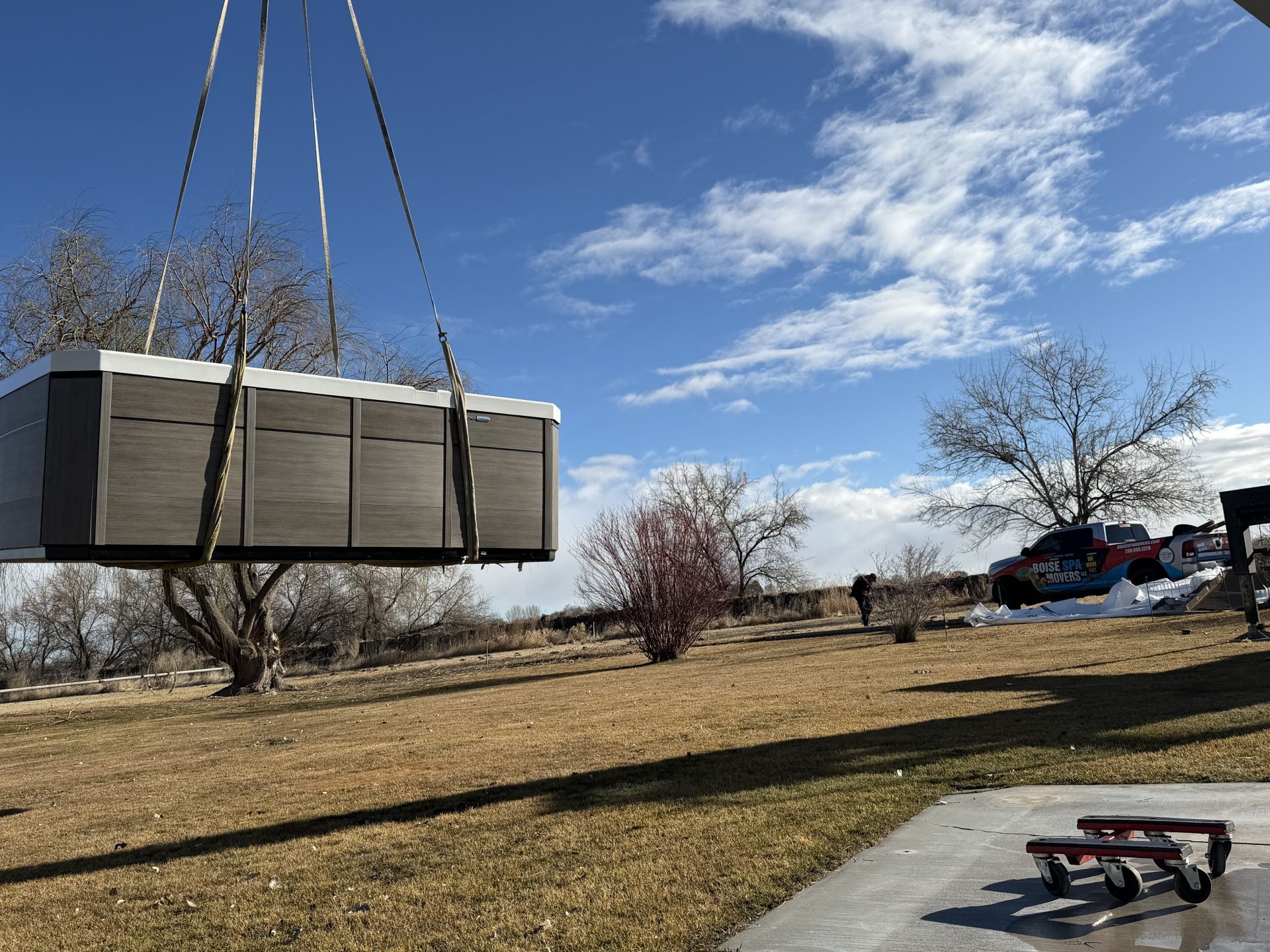 Hot tub being lifted by crane into a yard with brown grass and a blue sky.
