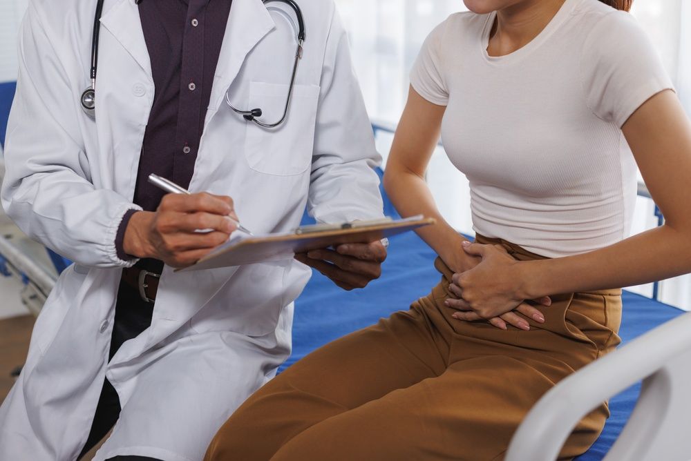 Doctor consulting a seated patient in a clinic, holding a clipboard and pen.