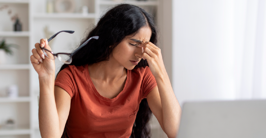 A woman is sitting at a desk with a laptop and rubbing her eyes.