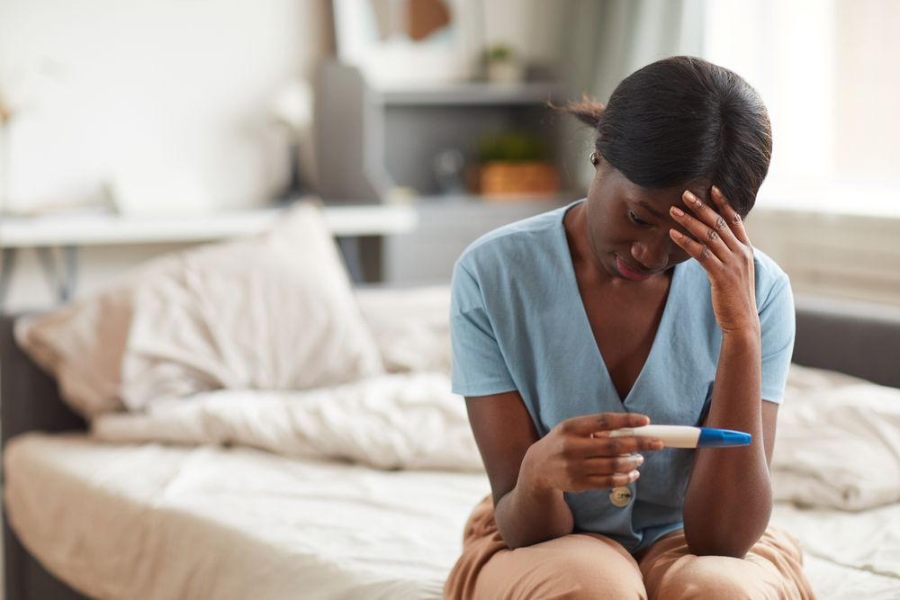 A worried young African-American woman looking at pregnancy test while sitting on bed at home.