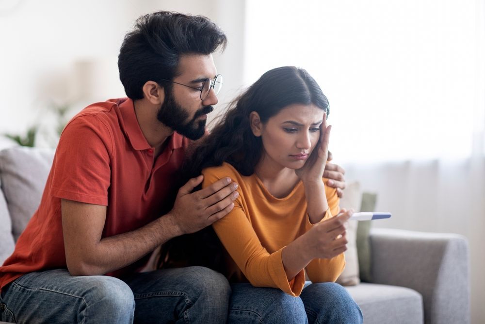 A young concerned woman looking at a pregnancy test while her boyfriend counsels her.