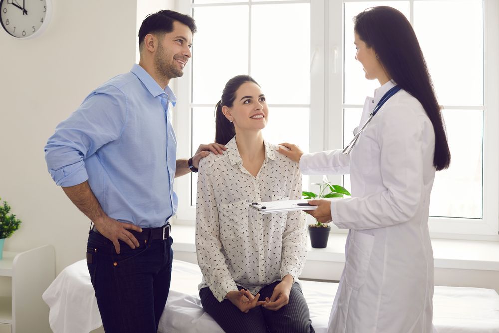 A doctor consults with a couple in an office. The woman is seated, and the man has his arm around her.
