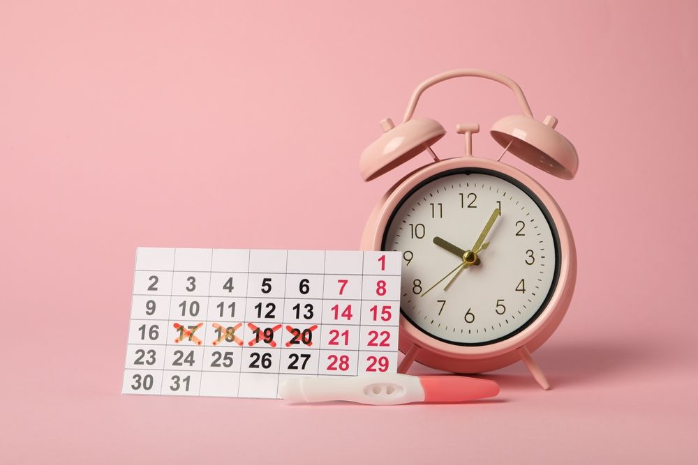 Calendar with crossed-out dates, a pink alarm clock, and a pregnancy test on a pink background.