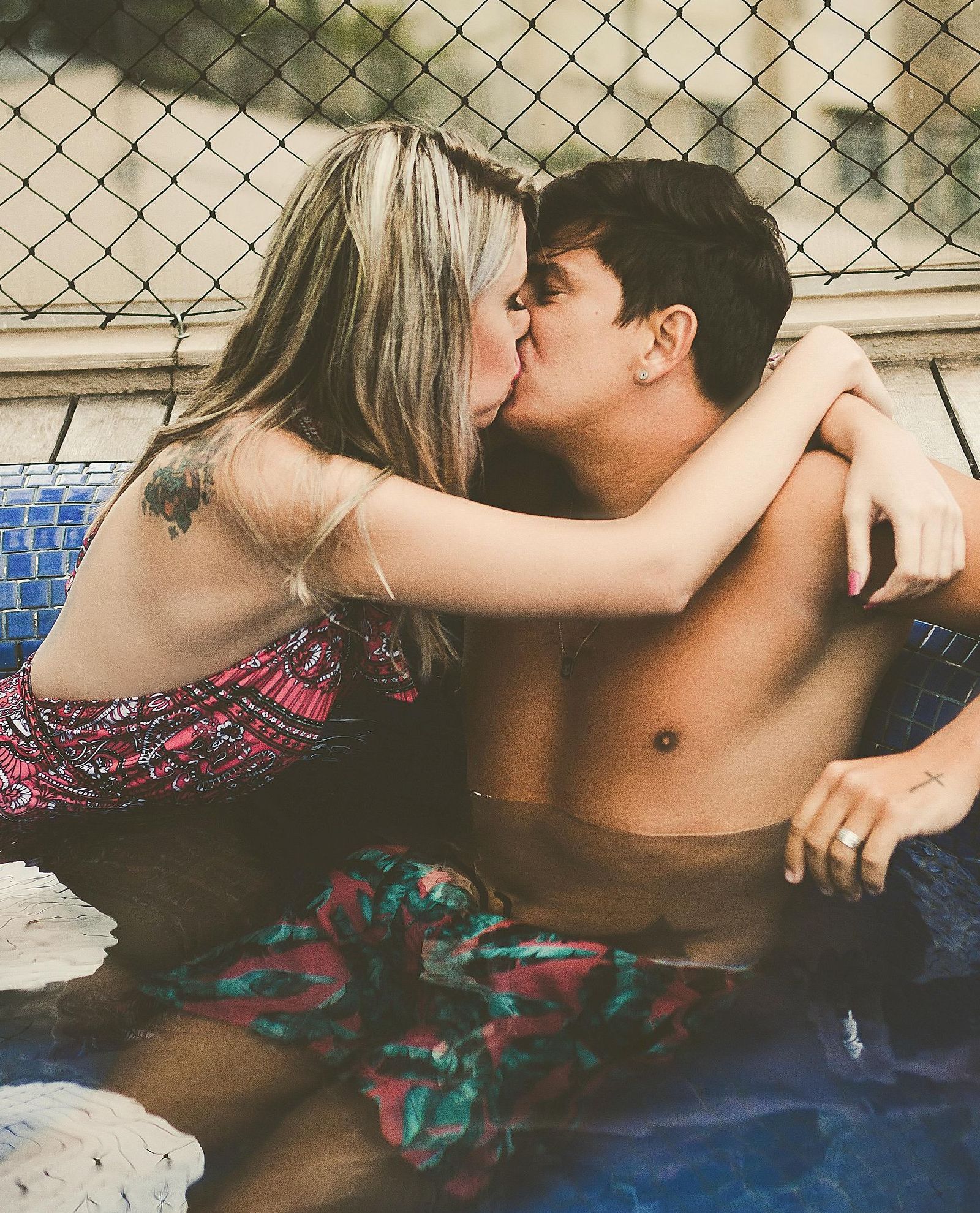Couple kissing in a blue hot tub; woman in pink dress, man shirtless. City backdrop.