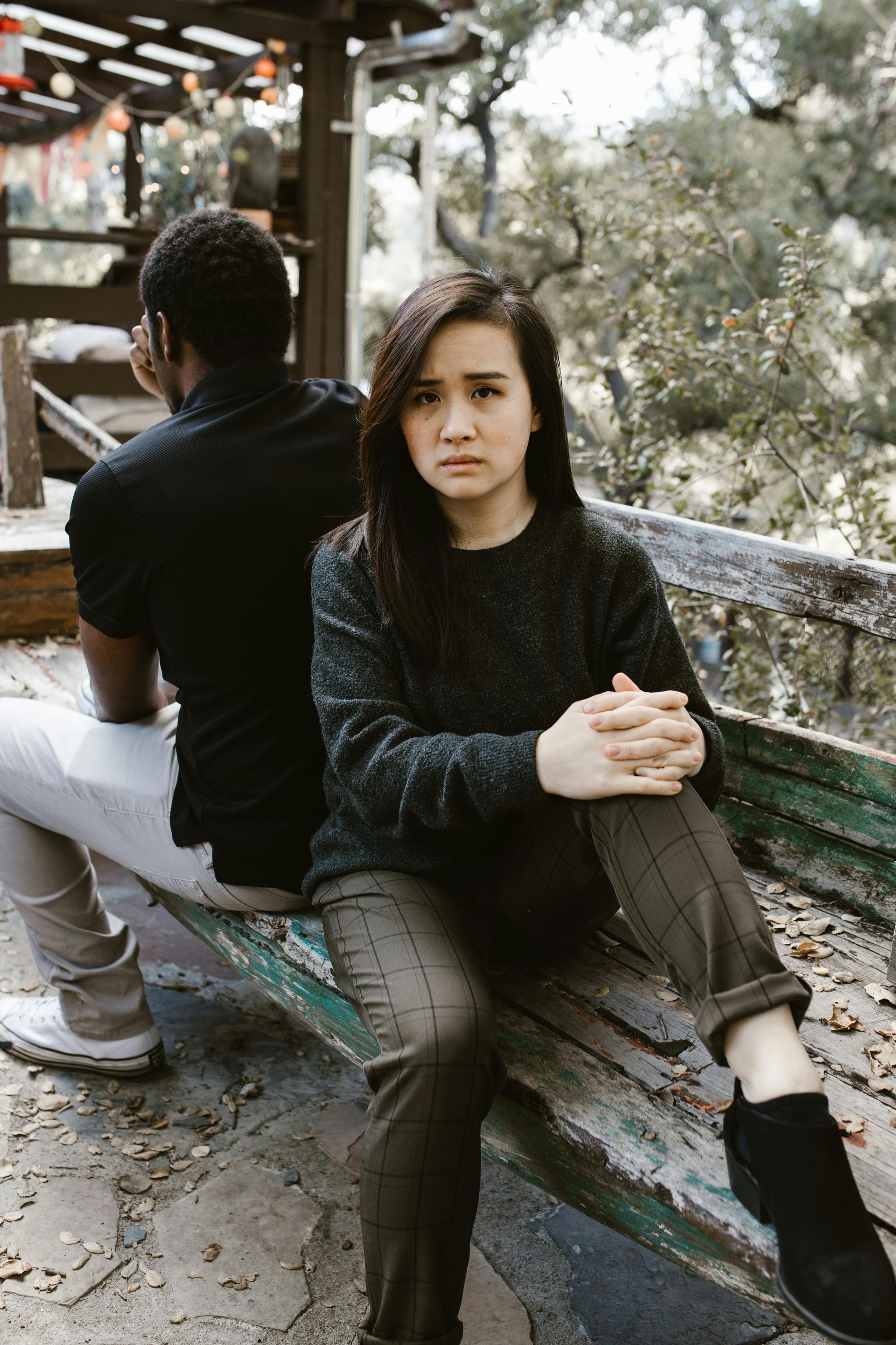 Woman with serious expression sits next to a man turned away, on a weathered bench outdoors.