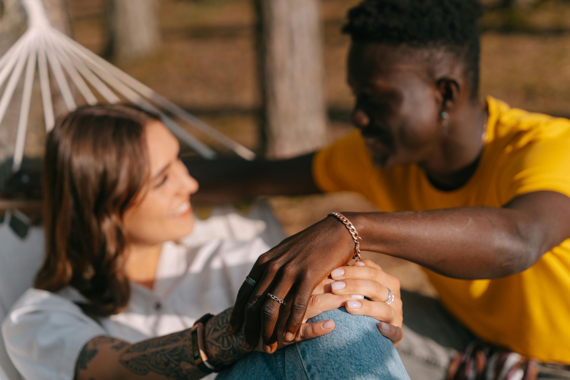 Woman in hammock gazes at smiling Black man, who touches her hands. They are outdoors.