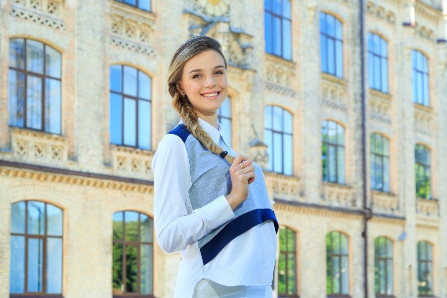 A young woman is standing in front of a large building holding a book.