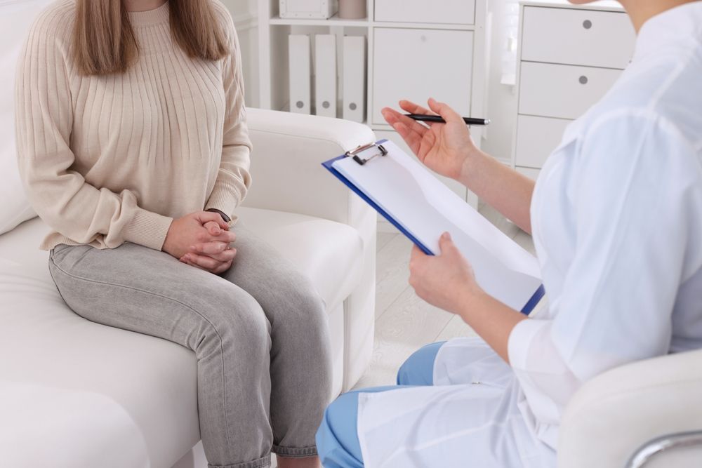 A woman is sitting on a couch talking to a doctor who is holding a clipboard.