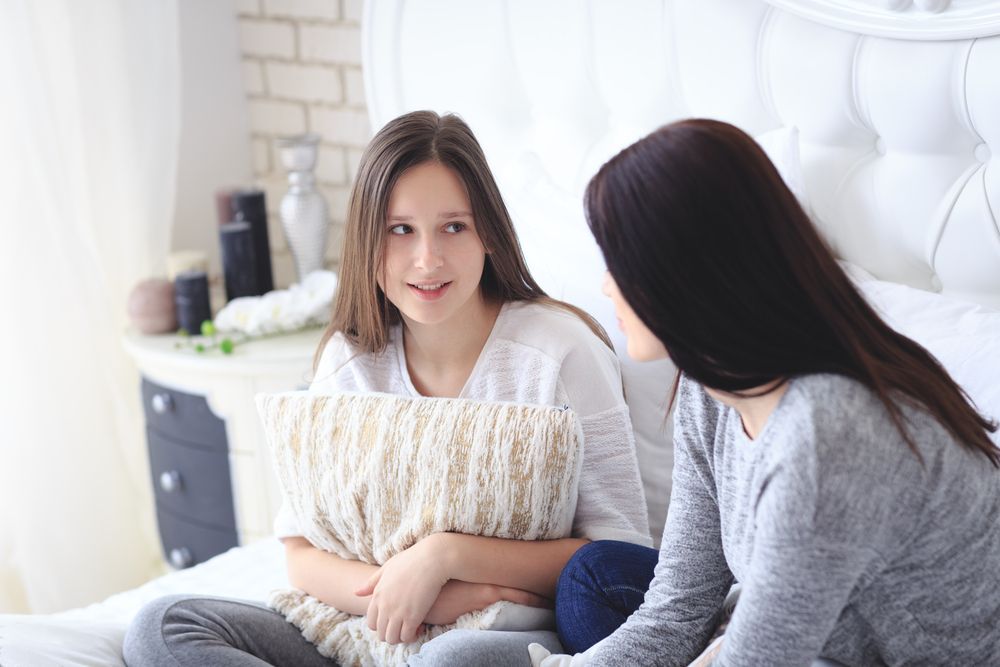 Two young women are sitting on a bed talking to each other.