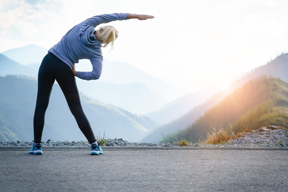 A woman is stretching her legs on a road in front of a mountain.