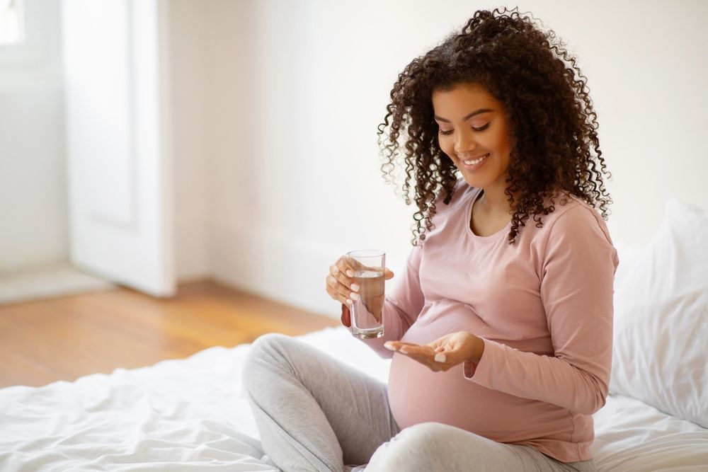 A pregnant woman is sitting on a bed taking a prenatal vitamins and drinking water.