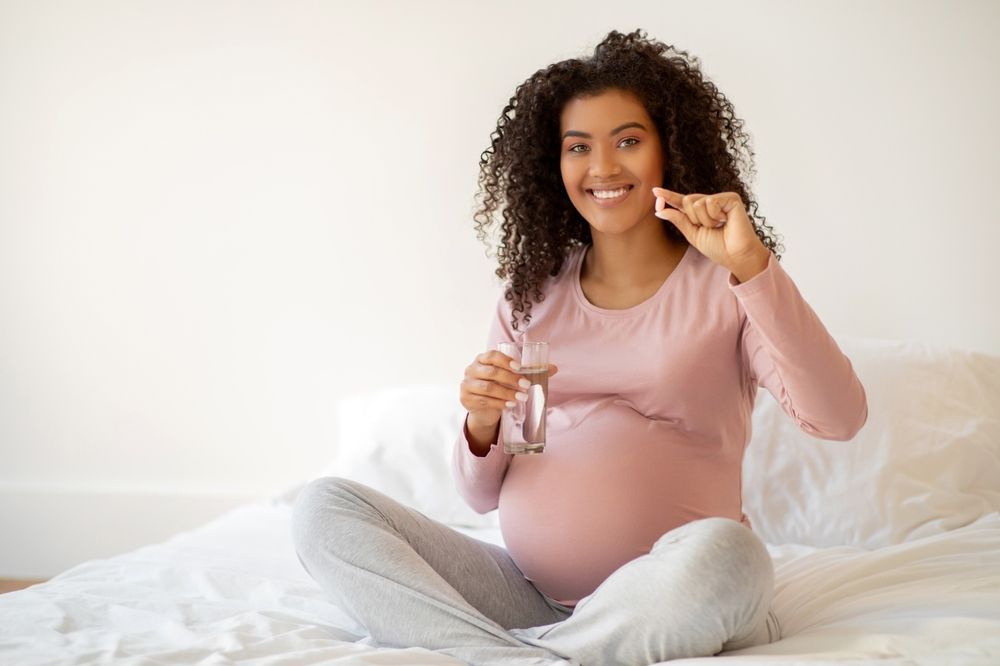 A pregnant woman is sitting on a bed holding a glass of water and a pill.