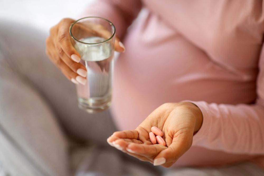 A person holding a glass of water and pink pills, with their pregnant belly visible.
