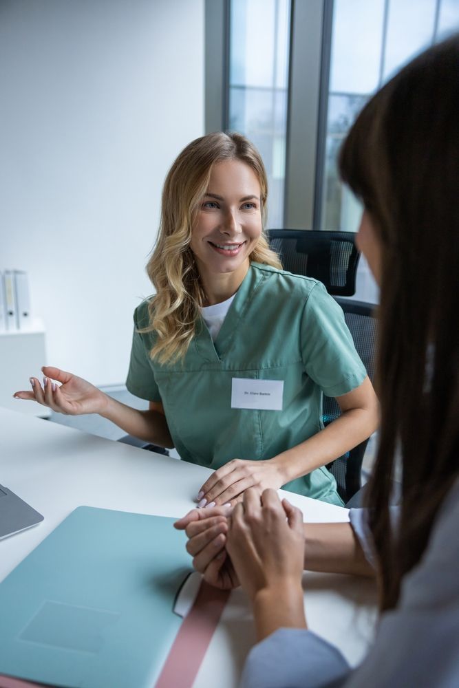 Nurse in green scrubs smiles, gesturing, talking to a patient at a desk in an office setting.