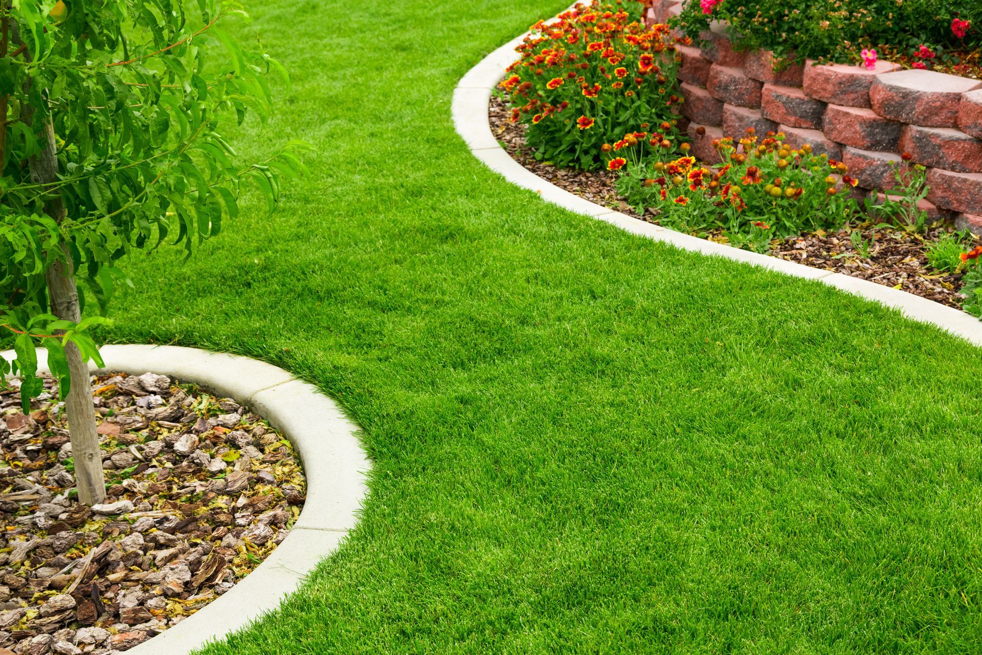 Curved garden path edged with white stone borders, green grass, flowers, and a brick retaining wall