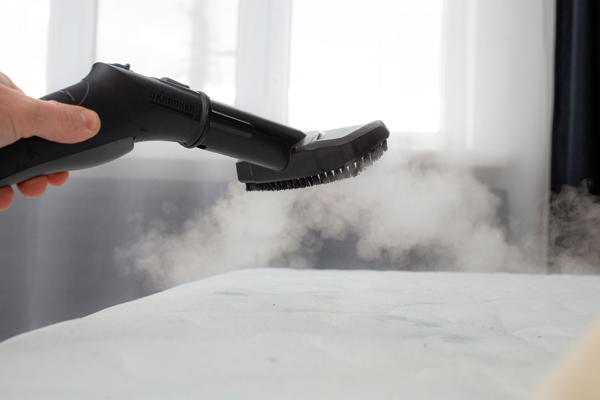 A person uses a handheld steam cleaner on a white surface, emitting visible steam in a room.