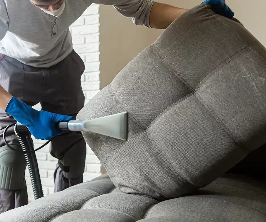 Person cleaning a gray couch with a handheld upholstery cleaner, wearing gloves.