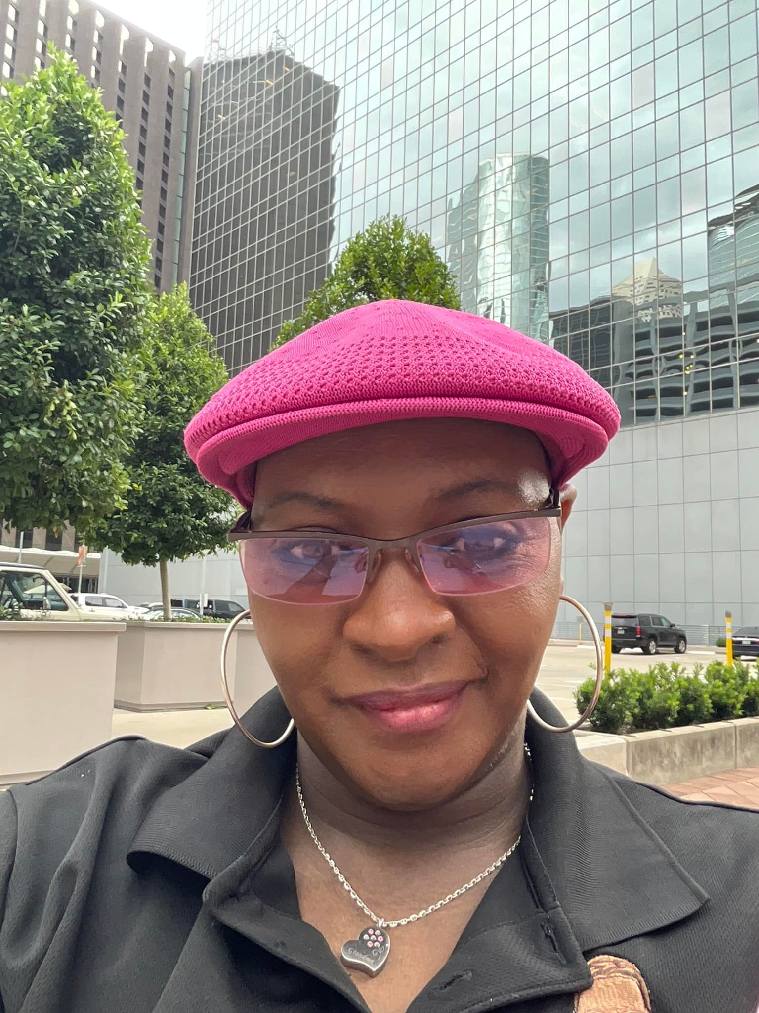 A woman wearing a pink hat and glasses is standing in front of a building.