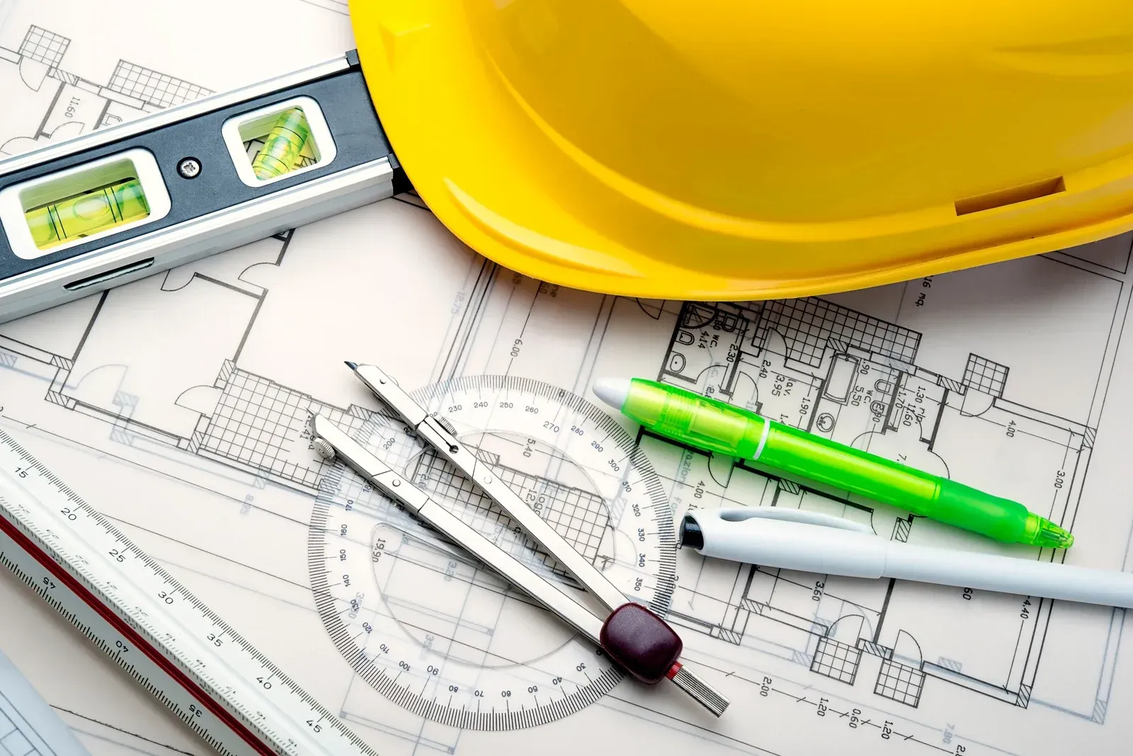 Yellow hard hat, blueprints, and tools for construction, including level, pen, and compass.