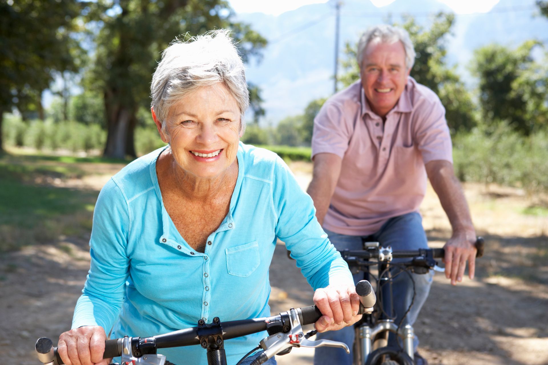 Two cyclists smiling on a sunny dirt path, riding outdoors with trees in the background