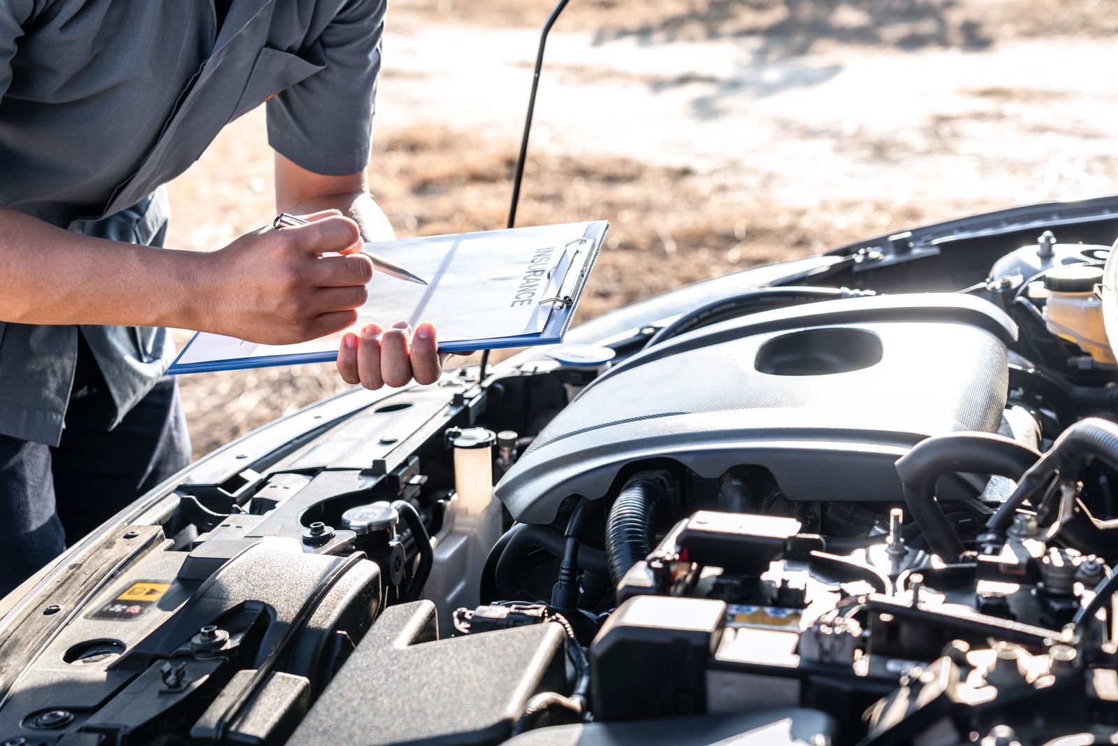 Mechanic checking a car engine, holding a clipboard and pen outdoors.