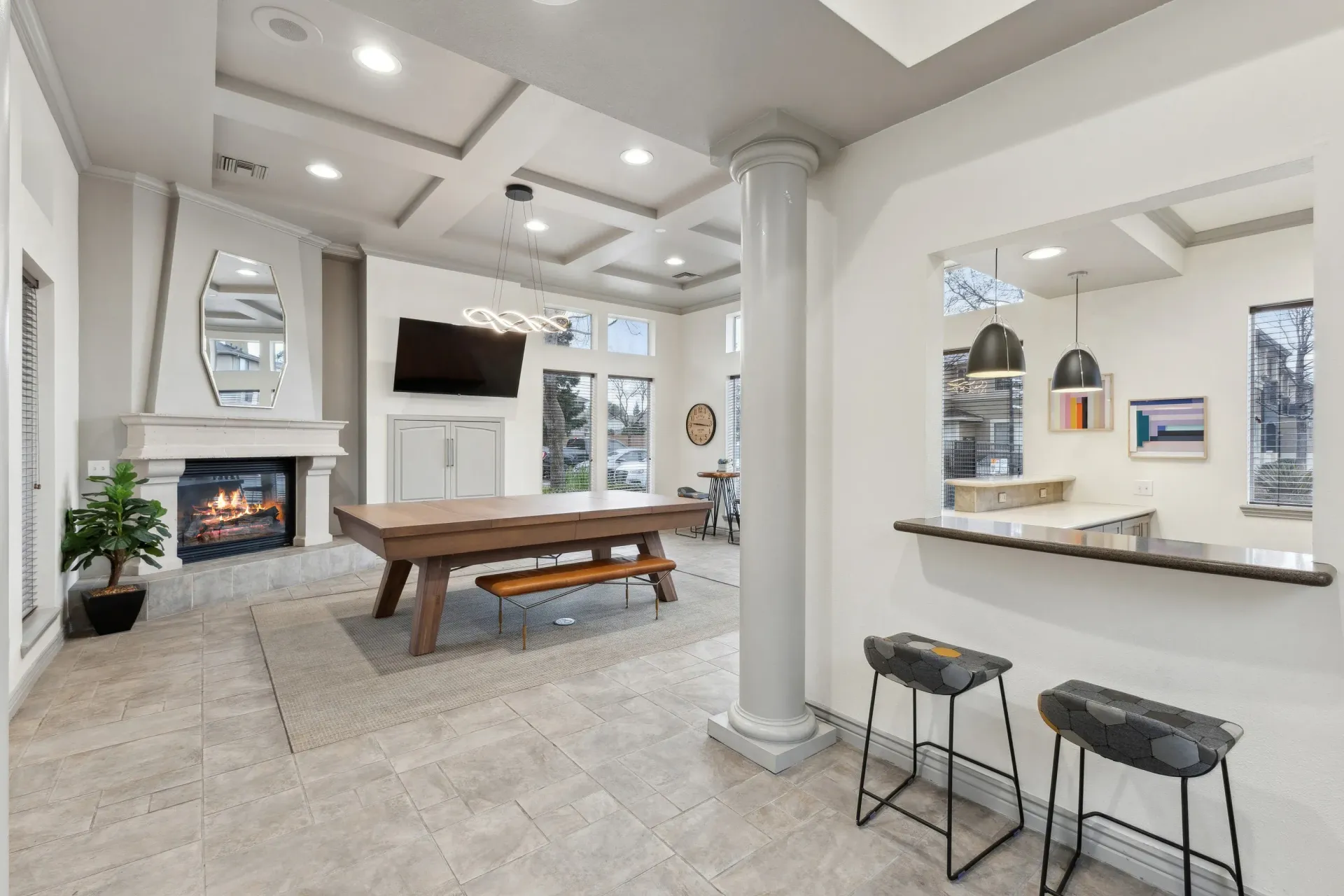 Bright community lounge with a fireplace, large wooden table, mounted TV, and bar seating.